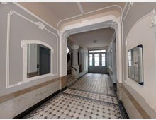 Eingangsbereich eines Altbaus mit dekorativen Wandverzierungen in Weiß und grauen Akzentfarben. Der Boden ist mit einem geometrisch gemusterten Fliesenmosaik gestaltet, und im Hintergrund befinden sich eine doppelflügelige Glastür sowie eine Treppe.