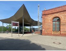 Bahnhofsvorplatz mit einem modernen überdachten Eingangsbereich, der durch rautenförmige Säulen gestützt wird, links ein digitaler Anzeigebildschirm und rechts ein historisches Backsteingebäude mit einem großen Rundbogenfenster. Im Hintergrund sind Bäume und Eisenbahnschienen zu sehen.