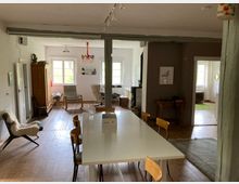 Offener Wohnbereich mit Holzboden, weißen Wänden und Balkendecke. Im Vordergrund steht ein großer weißer Esstisch, im Hintergrund sind Fenster, ein Holzschrank und Sitzmöbel erkennbar, die Zimmer durch breite Durchgänge verbunden.