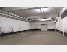 Ein leerer, industrieller Raum mit Betonboden und weißen Backsteinwänden, die teils von Schimmel und Verfärbungen gezeichnet sind. An der Decke befinden sich freiliegende Rohre und Leuchtstofflampen, eine Wand zeigt eine zugemauerte Türöffnung.