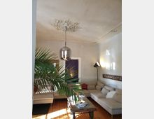 Wohnzimmer mit hellen Wänden und verziertem Stuck an der Decke, ausgestattet mit einer grauen Eckcouch, einem Holztisch auf Metallrädern und einer Stehlampe. Im Vordergrund steht eine Palme, und der Raum hat einen Holzboden sowie moderne und rustikale Designelemente.