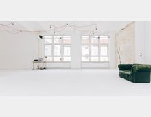 Heller, minimalistischer Raum mit weißen Wänden und einem Holzboden, großen Fenstern mit Blick auf Nachbargebäude, rohen Ziegeln an einer Wand, einem grünen Ledersofa auf der rechten Seite und einem kleinen Schreibtisch mit Laptop auf der linken Seite. Von der Decke hängen rote Kabel mit Glühbirnen.