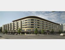 Ein sechsstöckiges modernes Gebäude mit geschwungener Fassade, gelblichen Kacheln und großen Fenstern in gleichmäßiger Anordnung. Das Gebäude steht an einer Straßenkreuzung mit Bäumen entlang des Gehwegs; im Erdgeschoss befinden sich Geschäfte, darüber ist ein Hotel.