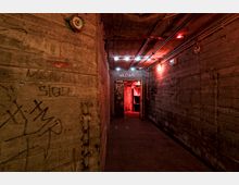 Betonflur in einem industriellen oder ehemaligen Bunkergebäude, beleuchtet von roten und weißen Lampen. Die Wände sind mit Graffiti bedeckt, und Rohre verlaufen entlang der Decke; durch eine Tür am Ende des Gangs ist ein rötlich beleuchteter Raum sichtbar.