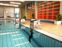 Hallenbad mit einem rechteckigen Schwimmbecken, Startblöcken entlang des Beckenrandes und einer farbenfrohen Kachelwand im Hintergrund. Der Bereich ist hell beleuchtet, mit Pflanzen und Sitzgelegenheiten an der Seite.