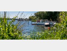 Boote an einem Steg auf einem See, umgeben von üppigem Schilf und Bäumen am Ufer. Im Hintergrund sind weitere Wasserfahrzeuge und eine grüne Uferlandschaft zu sehen.