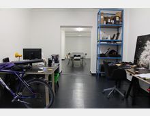 Ein schlicht eingerichtetes Büro mit einem schwarzen Fliesenboden, weißen Wänden und mehreren Arbeitsplätzen. Im Vordergrund stehen Schreibtische mit einem Computer, einem Regal, einem Fahrrad und Büromaterialien; im Hintergrund ist ein Besprechungstisch mit Stühlen zu sehen, umgeben von einer Schiebetür.