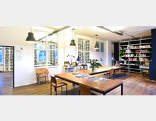 Ein heller, offener Arbeitsraum mit Holzfußboden, großen Holztischen und modernen Hängelampen. Die Wände sind mit Regalen voller Bücher und Pflanzen dekoriert, und auf einem langen Tisch stehen Schreibtischlampen sowie ein Blumenstrauß; ein Nebenraum ist durch Fenster und eine Tür erkennbar, die mit asiatischen Schriftzeichen verziert sind.