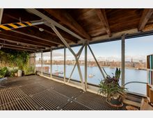 Innenraum eines Gebäudes mit Gitterboden, Holzbalkendecke und großen Fensterfronten, die den Blick auf einen Fluss mit Booten und umliegende moderne Gebäude ermöglichen; am Fenster und an der Wand stehen zahlreiche Pflanzen.