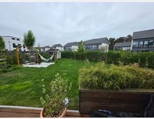 Blick über einen gepflegten Garten mit großer Rasenfläche, Hecken und jungen Bäumen in einem Wohngebiet mit modernen Einfamilienhäusern im Hintergrund. Links steht eine Holzpergola mit Hängesesseln und Hängematte auf einer Kiesfläche neben einem kleinen Gartenhaus.