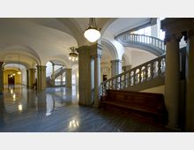 Großzügige Eingangshalle eines historischen Gebäudes mit glänzendem Marmorboden, massiven Säulen, geschwungener Treppe mit Balustrade und dekorativen Deckenleuchten. Tageslicht fällt durch große Fenster in den Raum.