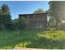 Blick auf eine große, verwitterte Scheune aus Holz mit wenigen Fenstern, umgeben von einer Wiese mit hohem Gras und einzelnen Gartenobjekten. Rechts stehen unter einem großen Baum Sitzmöbel im Freien, im Hintergrund weitere Bäume unter klarem Himmel.