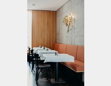 Ein modernes Restaurant mit einer Sitzbank an einer Betonwand, dekoriert mit einer Metall-Wandverzierung in Form von Blättern. Die Tische sind mit weißen Tischdecken bedeckt und mit schlichten Gläsern und Besteck gedeckt; schwarze Holzstühle ergänzen das Interieur.