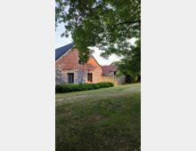 Ein ländliches Gebäude aus Naturstein und roten Ziegeln mit einem schwarzen Schrägdach, umgeben von einer Grünfläche und dichten Bäumen im Vordergrund. Im Hintergrund ist ein Holzzaun und ein weiteres Gebäude mit rotem Dach sichtbar.