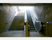 Treppenaufgang und Rolltreppe in einem U-Bahnhof mit modernen, glatten Wänden aus grauem Stein. Oben ist ein heller Ausgang erkennbar, der ins Freie führt.