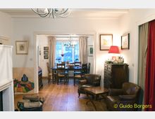 Wohnzimmer mit Holzboden und gemütlicher Einrichtung, im Vordergrund Ledersessel und ein kleiner Tisch, im Hintergrund ein Essbereich mit Holztisch und Stühlen vor einem Fenster mit Vorhängen. Eine Kommode mit Lampe und Dekorationen steht an der Seite, und ein Kronleuchter hängt von der Decke.