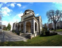 Historische Kapelle mit einer Kuppel und Säulenarkaden, umgeben von einem gepflegten Garten mit Grasflächen, Bäumen und einem gepflasterten Weg; im Hintergrund sind Wohngebäude zu sehen.