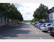 Straße mit geparkten Autos auf beiden Seiten, flankiert von kleinen Gebäuden und schattenspendenden Bäumen. Am Ende der Straße ist ein See mit bewaldetem Ufer im Hintergrund zu sehen.