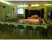 Ein Festsaal mit Holzboden, in dem lange, mit weißen Tischdecken und rot-weißen Servietten dekorierte Tische in U-Form aufgestellt sind. Im Hintergrund befinden sich eine kleine Bühne mit Rednerpult und Lautsprechern sowie ein an der Decke montierter Beamer und ein projiziertes Bild.