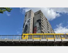 Wohnhochhaus mit grauer Fassade und großflächigen Wandgemälden, das im Vordergrund von einer erhöhten Bahntrasse mit einer gelben U-Bahn überspannt wird; blauer Himmel und Baumzweige im Hintergrund.