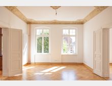 Heller Raum mit hohen Decken, großen Fenstern und Zierleisten an der Decke. Der Boden besteht aus Parkett, und durch die Fenster ist der Blick auf Bäume und ein Gebäude mit Ziegeldach zu sehen.