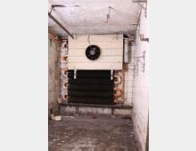 Ein leerer, verschmutzter Kellerraum mit einer an der Wand montierten alten Kühlanlage, bestehend aus einem Lüfter und schwarzen Rippenrohren. Die Wände sind mit weißen, teilweise beschädigten Fliesen verkleidet, und der Boden ist staubig und uneben.