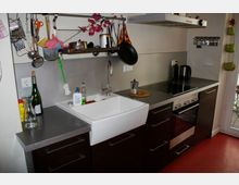 Moderne Küche mit dunklen Unterschränken, einer weißen Spüle und einer Arbeitsplatte aus Metall. Oben sind Regale mit Küchenutensilien, daneben ein Herd mit Dunstabzugshaube; eine Glastür führt nach draußen.