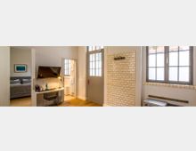 Helles Zimmer mit modernem Design, bestehend aus einem Arbeitsbereich mit Schreibtisch, Stuhl und Wandmonitor, einer angrenzenden Schlafnische mit grauem Bett und einem Badezimmer im Hintergrund. Die Wände weisen teils cremefarbene Fliesen auf, die Fenster haben große Gitterrahmen.