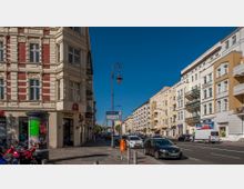 Straßenszene mit Altbauten, darunter ein rot-weißes Gebäude mit einer Apotheke im Erdgeschoss sowie weiteren Wohnhäusern mit Balkonen rechts im Bild. Im Vordergrund befinden sich ein Gehweg, Straßenschilder, eine Laterne und parkende Autos auf der rechten Seite der Fahrbahn.