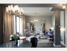 Elegantes Wohnzimmer mit modernen und klassischen Elementen, darunter ein großer Kronleuchter, ein ovaler Esstisch mit Blumenarrangement und gepolsterte Möbel in sanften Farben. Die Wände sind teils mit Tapeten in floralem Muster gestaltet, und große Fenster lassen natürliches Licht in den Raum.