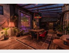 Gemütlicher Innenraum mit industriellem Design; Holzboden, offene Backsteinwände und große Fenster. Der Raum ist mit Sofas, einem Holztisch, Stühlen in verschiedenen Farben, Pflanzen sowie einer bunten Hängelampe und warmem Licht dekoriert.