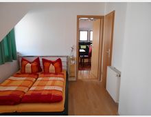 Schlafzimmer mit einem Doppelbett, das mit rot-orange kariertem Bettzeug bezogen ist, links ein Fenster mit grünem Vorhang. Rechts führt eine Tür in einen angrenzenden Raum, der ein Essbereich mit Tisch und Stühlen zeigt. Holzboden und weiße Wände prägen den Raum.