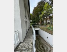 Außenbereich eines Gebäudes mit einem gepflasterten Gehweg, der von einem weißen Geländer begrenzt wird, neben einer Abfahrt zu einer Tiefgarage. Im Hintergrund sind Bäume und angrenzende Wohnhäuser sichtbar.