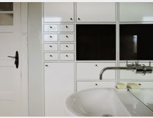 Weißes Badezimmer mit einem wandhängenden Waschbecken, einem modernen verchromten Wasserhahn und zwei Seifenstücken in Seifenschalen. Im Hintergrund ein weißer Wandschrank mit mehreren kleinen Schubladen und schwarzen Glaselementen, daneben eine weiße Tür mit schwarzem Türgriff.