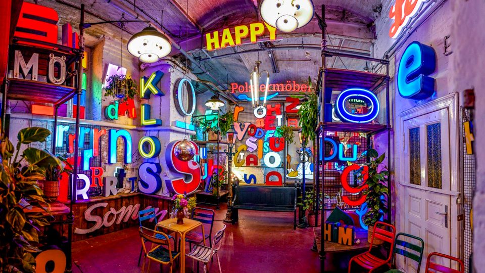 Innenraum eines bunt beleuchteten Cafés oder einer Bar in einem industriellen Gewölberaum mit sichtbaren Leitungen, Hängelampen und zahlreichen Neonbuchstaben an den Wänden. Im Raum stehen kleine Tische und Stühle, Pflanzen sowie metallene Regale mit Leuchtschildern.