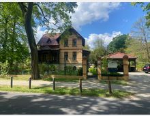 Historische Villa mit gelber Klinkerfassade und rotem Ziegeldach, umgeben von einem Zaun mit Eingangstor, davor ein Gehweg und Rasenfläche; rechts im Bild ein kleiner Carport mit rotem Dach sowie parkende Autos. Im Hintergrund Bäume und blauer Himmel.