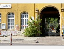 Gelbes Gebäude mit großen, weißen Sprossenfenstern und sichtbaren Graffiti-Spuren an der Fassade. Rechts befindet sich ein schmiedeeisernes Tor in einem Torbogen, das in einen Innenhof mit Bäumen und weiteren Strukturen führt.