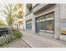 Straßenseite eines mehrstöckigen Wohn- und Geschäftshauses mit grauer Steinfassade, großen Schaufensterfenstern und einem Eingang unter einem abgerundeten Fassadenvorsprung. Vor dem Gebäude verläuft ein breiter Gehweg mit Straßenbäumen, Fahrradbügeln und geparkten Autos entlang der Allee.