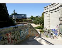 Blick auf eine urban gestaltete Treppe mit Graffiti an den Wänden, die zu einer Bahnstrecke führt, auf der eine gelb-rote S-Bahn zu sehen ist. Im Hintergrund befinden sich moderne Gebäude und Bäume unter einem klaren blauen Himmel.