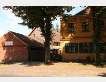 Backsteingebäude mit dem Schild „Lucke-Hof“, daneben ein gelbes, zweistöckiges Haus mit grünen Fensterläden, und im Hintergrund ein Fachwerkgebäude. Im Vordergrund stehen Bäume und eine gepflasterte Straße mit Gehweg.