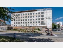 Eine unrenovierte weiße Gebäudefassade mit mehreren quadratischen Fenstern, bezeichnet als "City Hotel Lunik", flankiert von einer bepflanzten Verkehrsinsel im Vordergrund. Im Bild fährt ein Rollerfahrer auf einer Straße vor dem Gebäude entlang, während im Hintergrund weitere Häuser und Bäume sichtbar sind.