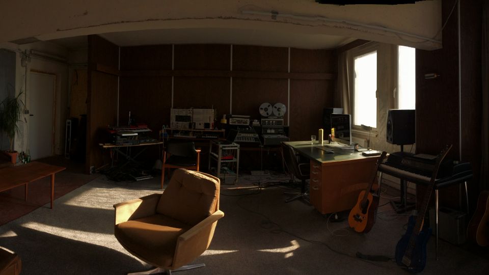 Panoramablick in einen großzügigen, loftartigen Raum mit abgenutzten Wänden, großen Fenstern mit Blick auf Hochhäuser und einer Mischung aus Wohn- und Musikstudioeinrichtung. Zu sehen sind Sofa und Couchtische, ein Arbeitsbereich mit Technik sowie mehrere Gitarren und Verstärker entlang der Fensterfront.