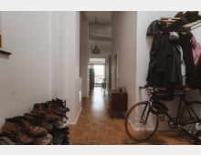 Schmaler Flur in einer Wohnung mit hellem Holzboden, links ein Schuhregal mit zahlreichen Schuhen, rechts eine Garderobe mit Jacken und einem Fahrrad. Am Ende des Flurs gibt es einen kleinen Schrank, eine Hängelampe und einen Blick auf eine gläserne Tür, die auf einen Balkon führt.