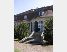 Historisches Gebäude mit ovaler Fensterfront und geschwungenem Ziegeldach, flankiert von hohen Bäumen. Eine steinerne Treppe führt zum Eingang mit hölzernen Fensterläden und Glastüren.