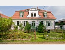 Ein zweistöckiges Wohnhaus mit Satteldach und roten Dachziegeln, grünen Fensterläden und einer verzierten Dachgaube. Vor dem Haus befindet sich ein gepflegter Garten mit Pflanzen, kleinen Bäumen und einem schmiedeeisernen Zaun.