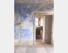 Eine Innenszene mit einer abblätternden, blau gestrichenen Wand, durchbrochen von einer offenen Tür mit schmalem Holzrahmen. Im Hintergrund ist ein weiterer Raum mit einem Fenster, Tageslicht und Möbeln wie einem grünen Sessel und Teppich sichtbar.