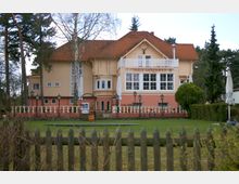 Zweistöckiges Gebäude mit orangefarbenem Anstrich und einem roten Ziegeldach, beschriftet mit "Haus Sanssouci". Vor dem Haus befinden sich eine Terrasse mit Laternen und eine gepflegte Grünfläche, umgeben von Bäumen und einem Holzzaun.