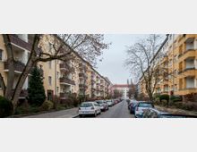 Straßenansicht einer Wohngegend mit mehrstöckigen, gelb verputzten Gebäuden und rotbraunen Klinkerelementen. Links und rechts der Straße sind parkende Autos sowie Bäume ohne Laub sichtbar; im Hintergrund erstrecken sich weitere Wohnhäuser.