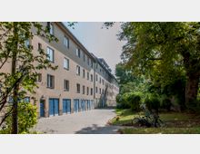 Mehrstöckiges Wohngebäude mit beigen Fassaden und blauen Garagentoren vor einem gepflasterten Weg. Rechts befindet sich eine Grünfläche mit Bäumen und zwei abgestellten Fahrrädern.