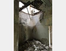 Ein verlassenes, stark beschädigtes Zimmer mit eingestürztem Dach, freiliegenden Holzträgern und Schutthaufen auf dem Boden. Die Wände sind abgenutzt und teilweise mit abblätternder Farbe bedeckt; rechts ist eine geöffnete Tür sichtbar.